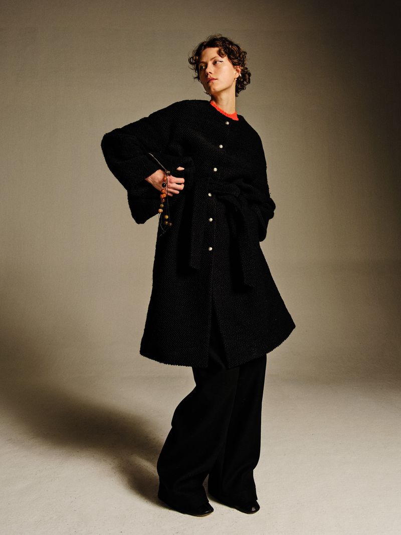 Model wearing a black belted button-up coat and matching wide-leg trousers against a warm studio backdrop.