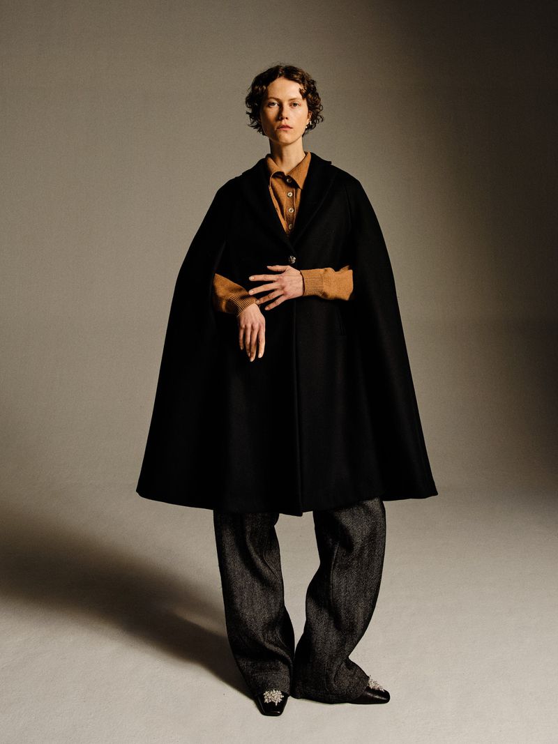 A young woman poses indoors wearing a black cape, brown collared shirt, and gray pants against a neutral background, fashion studio lighting.