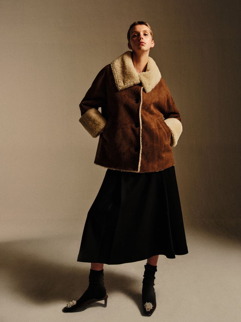 A fashion shot of a woman in a brown shearling coat and black skirt, posing in a studio with soft lighting, showcasing an elegant winter look.