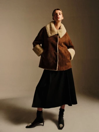A fashion shot of a woman in a brown shearling coat and black skirt, posing in a studio with soft lighting, showcasing an elegant winter look.