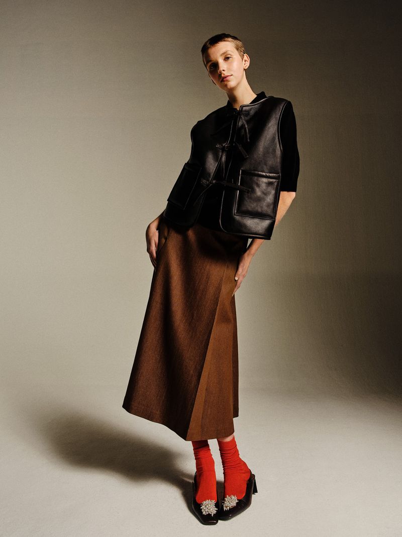 Full-length studio shot of a young woman wearing a black leather vest, brown skirt, and red socks, posing against a gradient gray background.