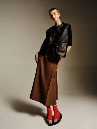 Full-length studio shot of a young woman wearing a black leather vest, brown skirt, and red socks, posing against a gradient gray background.