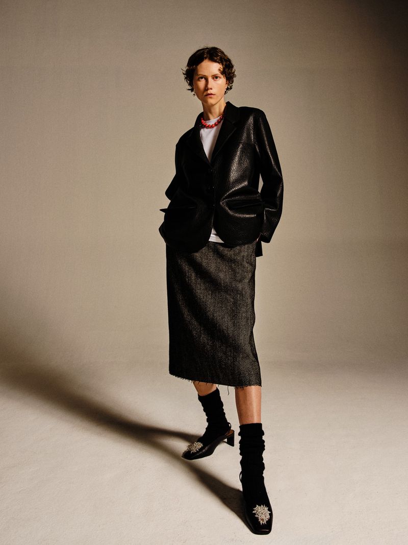 Adult female wearing a shiny black blazer, gray skirt, and black heeled shoes in a studio, standing and looking at the camera.