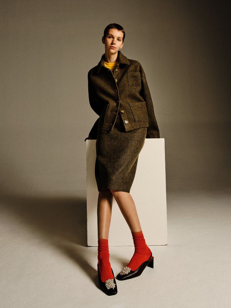 Model Bente Oort posing in an olive wool skirt suit, bright red socks, and black embellished heels against a studio backdrop.