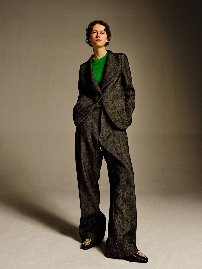 Full-length portrait of a woman in a gray suit and green shirt on a gray background, presented in a studio setting with soft, diffused light.