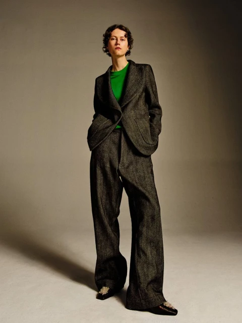 Full-length portrait of a woman in a gray suit and green shirt on a gray background, presented in a studio setting with soft, diffused light.