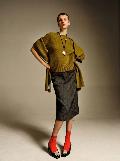 Full studio shot of a young adult woman in a green sweater and grey skirt posing with her hands on her hips, accessorized with jewelry.