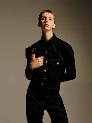 Model Bente Oort in a black button-down shirt with gold buttons and matching trousers holding a clutch for Rika Studios AW24 Lookbook.