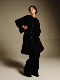 Model wearing a black belted button-up coat and matching wide-leg trousers against a warm studio backdrop.