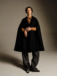 Model in a structured black cape coat, tan buttoned shirt, and charcoal wide-leg trousers posing against a grey studio backdrop.