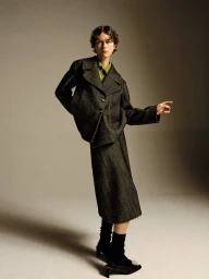 Model in dark wool blazer and matching skirt wearing black heels against a grey studio backdrop