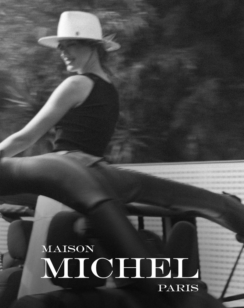 Black and white fashion shot of a woman wearing a hat, boots, and vest sitting on a vehicle with a blurred background.