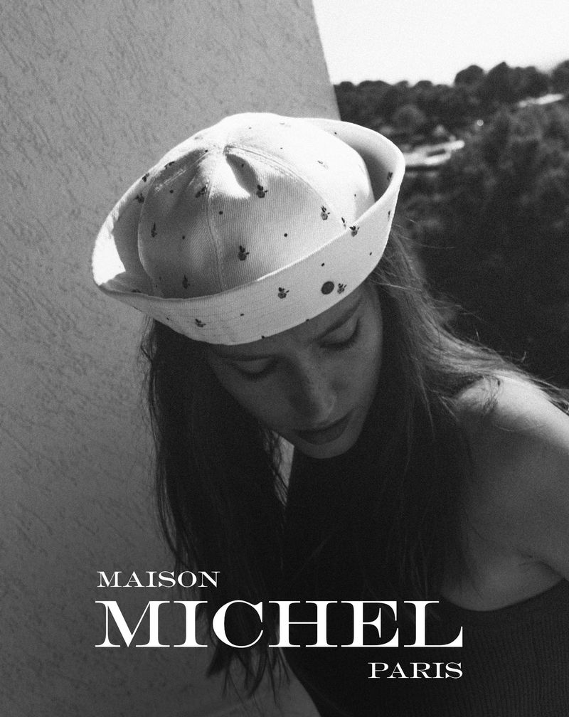 Black-and-white portrait of a woman wearing a white patterned Maison Michel sailor cap, posing outdoors with foliage in the background.