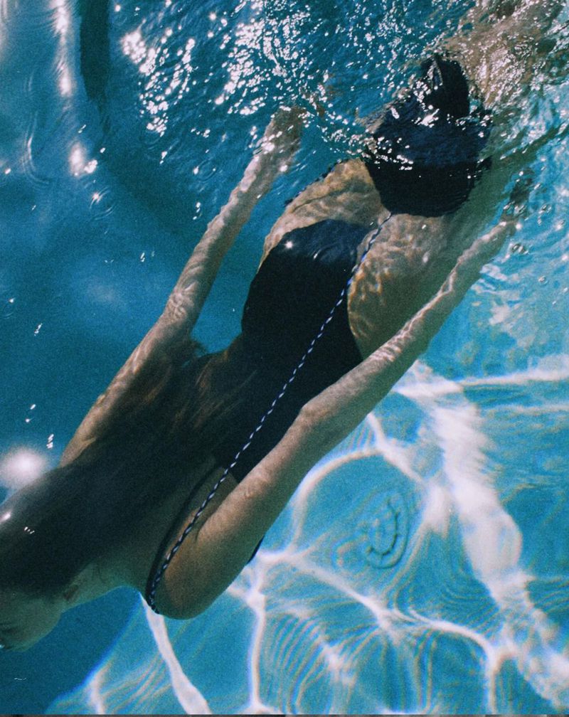 Underwater view of a woman swimming in a pool, with sunlight creating patterns on the pool's floor and sides.