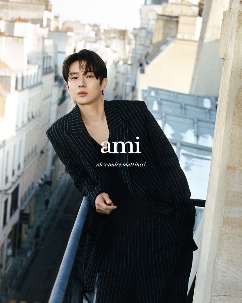 A fashionable Asian man poses on a city balcony in a pinstripe suit, with a backdrop of classic European architecture and natural light.