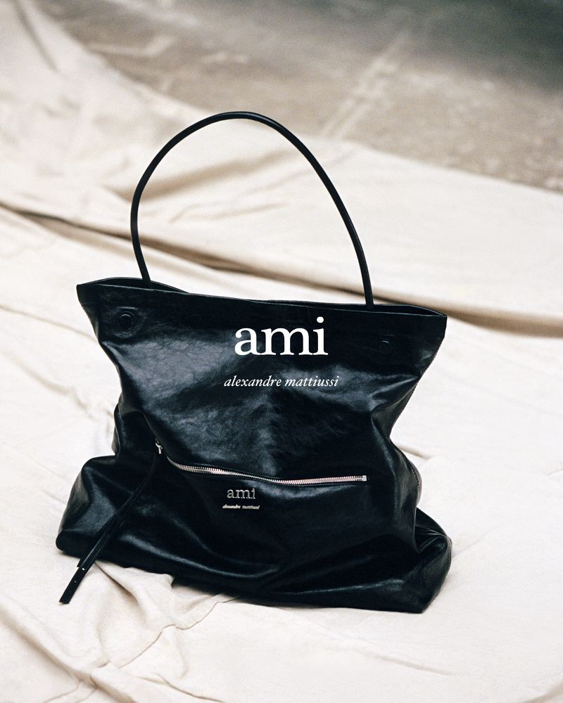 Black leather Ami handbag on off-white wrinkled fabric. Fashion product shot. Brand names include 'ami' and 'alexandre mattiussi'.