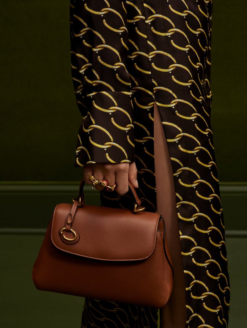 A brown leather handbag with a top handle is held by a person wearing a patterned dress and a statement ring, against a green backdrop.