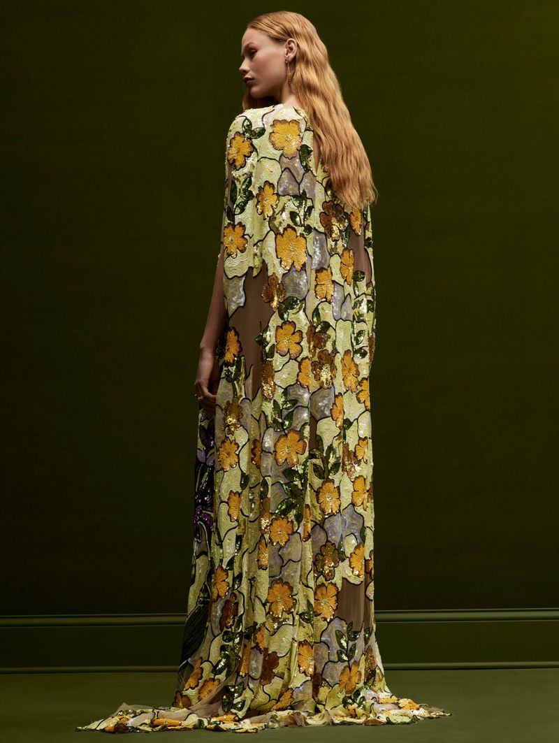 Woman in a long, floral-patterned gown with a cape poses in front of a dark green wall. Fashion, style, and beauty are the focus.