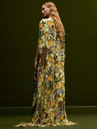 Woman in a long, floral-patterned gown with a cape poses in front of a dark green wall. Fashion, style, and beauty are the focus.