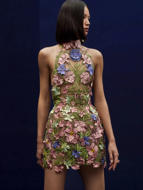 Adult Asian woman wearing floral halter dress against a dark blue background in a studio fashion shot; dress is made of pink and purple flowers.