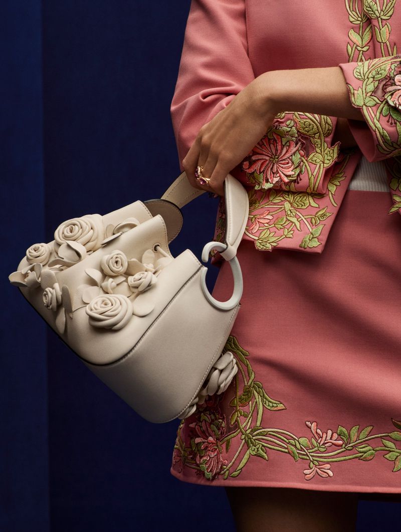 Close-up of a woman holding a cream-colored leather bucket bag adorned with white rose appliques against a dark blue background.