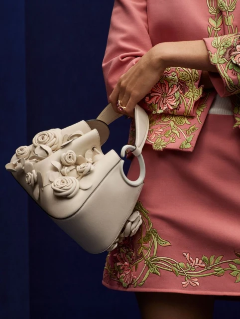 Close-up of a woman holding a cream-colored leather bucket bag adorned with white rose appliques against a dark blue background.