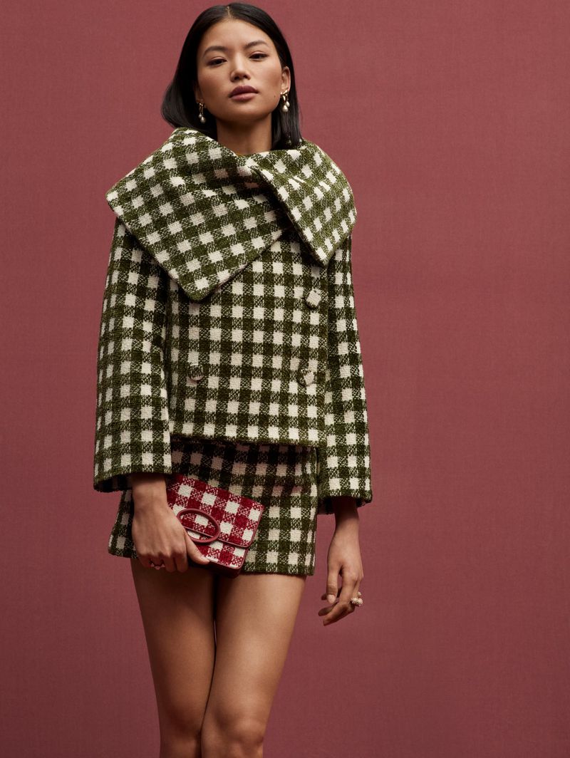 A woman poses in a green and white checked skirt suit with a small handbag against a solid burgundy backdrop in a studio setting.