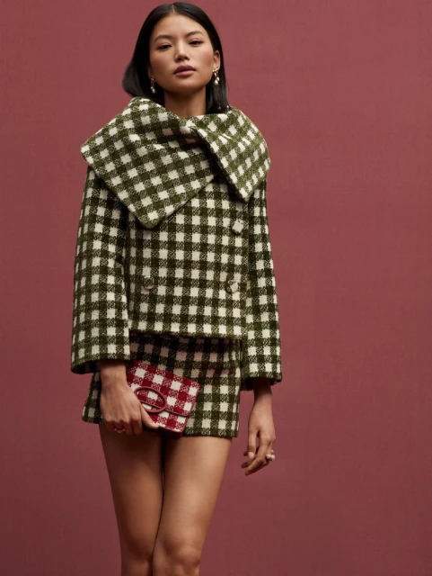 A woman poses in a green and white checked skirt suit with a small handbag against a solid burgundy backdrop in a studio setting.