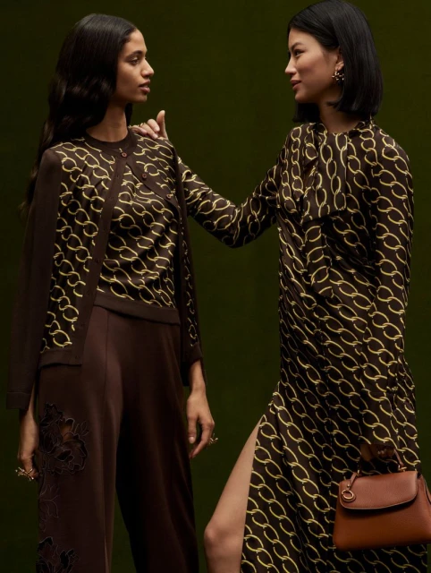 Two women in brown and gold patterned outfits pose against a dark green backdrop, one wearing a dress with a slit, and the other pants with lace.
