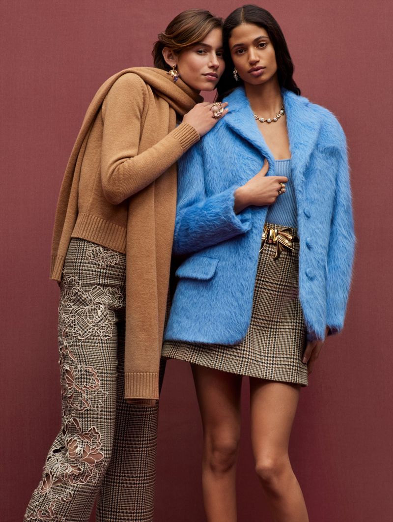 Two women pose in a fashion shot wearing a blue coat, scarf, statement jewelry, and patterned skirts on a maroon background.