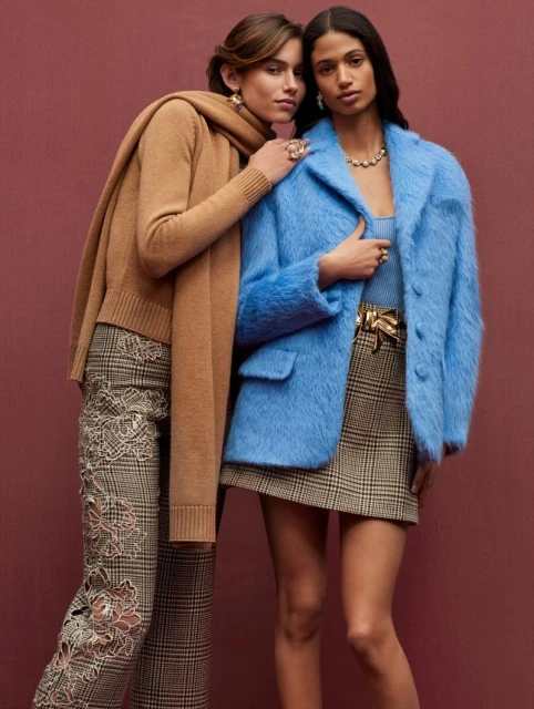 Two women pose in a fashion shot wearing a blue coat, scarf, statement jewelry, and patterned skirts on a maroon background.