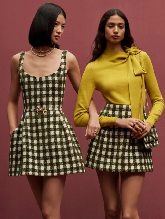 Two women in checkered dresses and skirt pose indoors. One woman wears a chartreuse sweater with a bow. Fashion editorial.