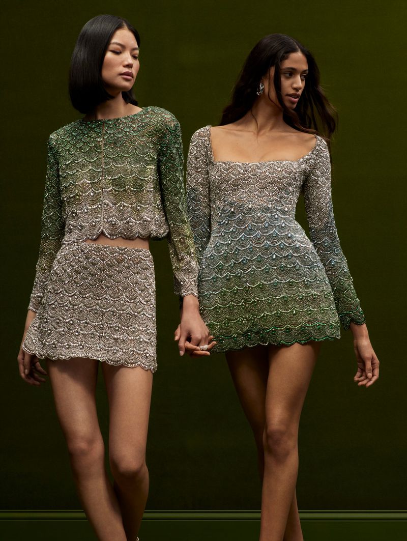 Two women pose in matching long-sleeve beaded mini dresses against a dark green backdrop, showcasing fashion and elegance.