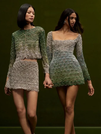 Two women pose in matching long-sleeve beaded mini dresses against a dark green backdrop, showcasing fashion and elegance.