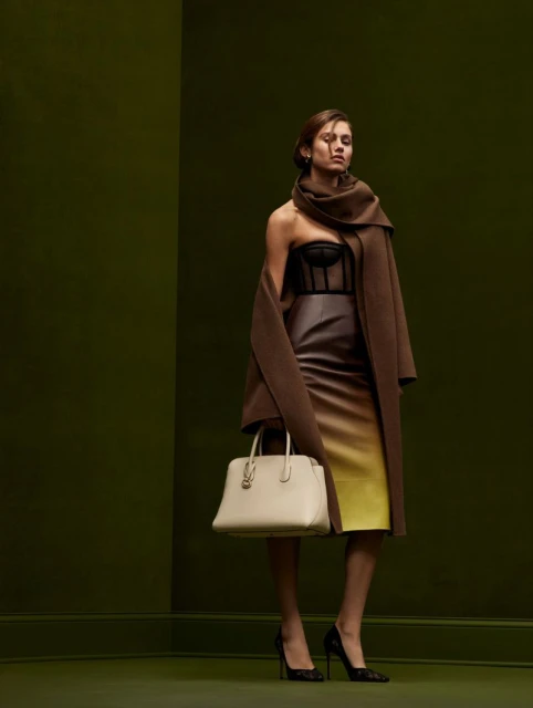 Adult woman in a brown coat, dress, and black high heels poses against a green studio background with a light-colored handbag.