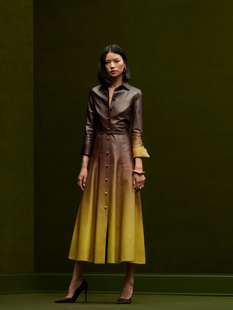 A full-length portrait of a young Asian woman in a brown and yellow ombre leather dress and brown heels against a green background.