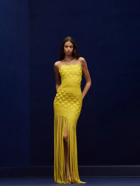 Full shot of a fashionable woman posing in a strapless yellow woven gown with fringe detail against a dark blue background.