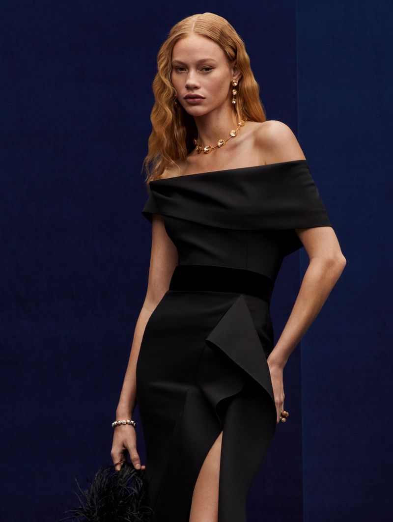 Elegant woman in off-the-shoulder black gown with slit, accessorized with gold jewelry, holding a feathered clutch against a dark blue backdrop.