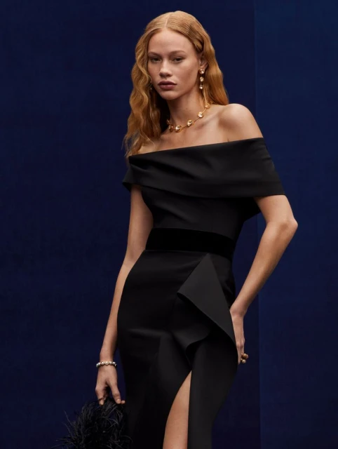 Elegant woman in off-the-shoulder black gown with slit, accessorized with gold jewelry, holding a feathered clutch against a dark blue backdrop.
