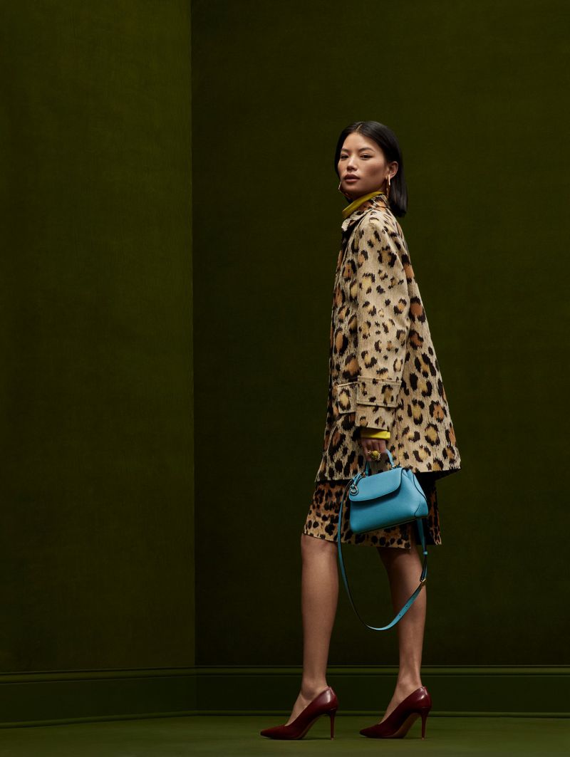 Asian woman in a leopard print coat and dress, accessorized with a blue handbag and maroon high heels, against a green backdrop.