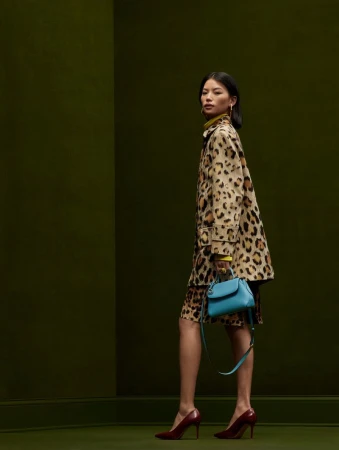 Asian woman in a leopard print coat and dress, accessorized with a blue handbag and maroon high heels, against a green backdrop.