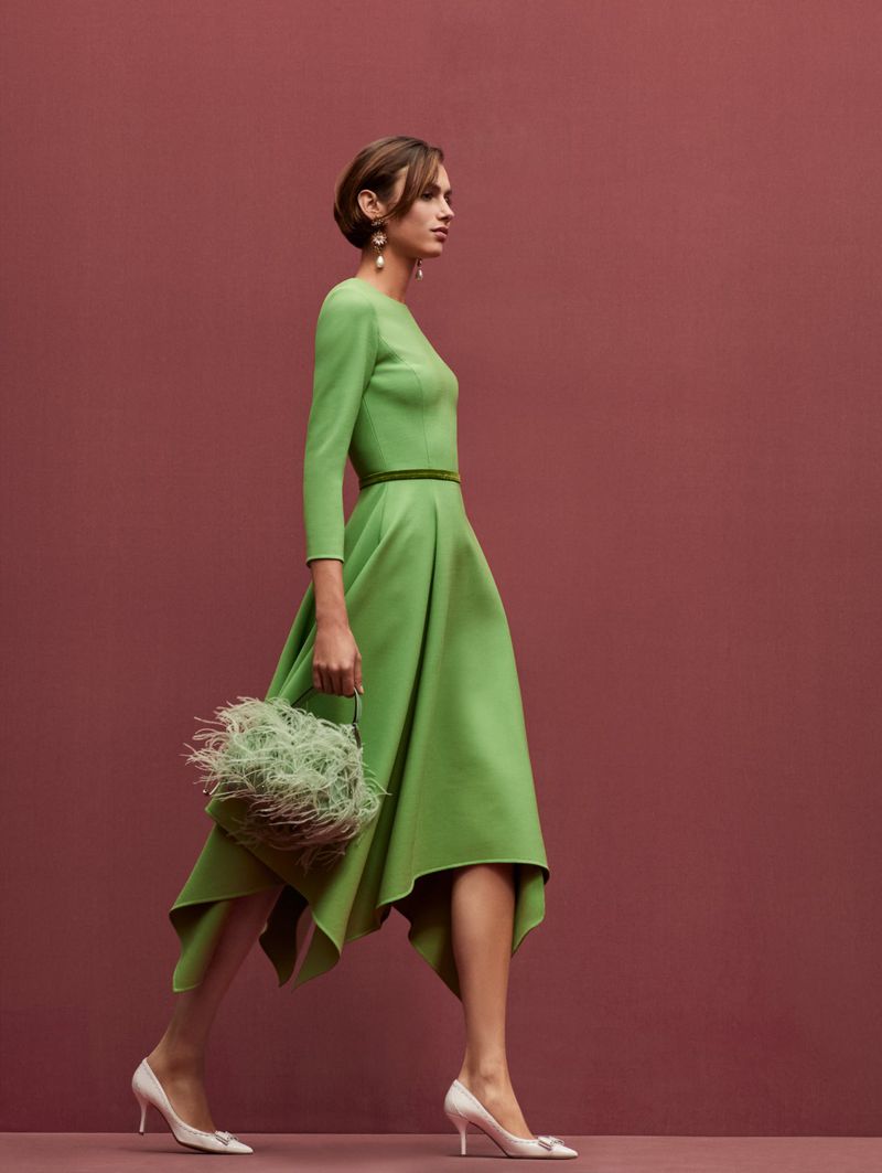 A woman in a green dress walks in profile against a mauve backdrop, carrying a feathered bag and wearing white high-heeled shoes.