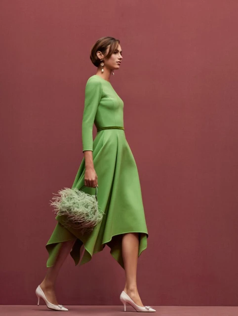 A woman in a green dress walks in profile against a mauve backdrop, carrying a feathered bag and wearing white high-heeled shoes.