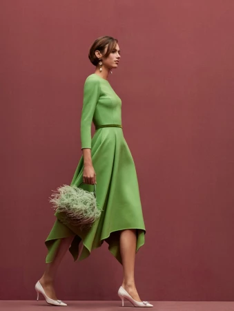 A woman in a green dress walks in profile against a mauve backdrop, carrying a feathered bag and wearing white high-heeled shoes.
