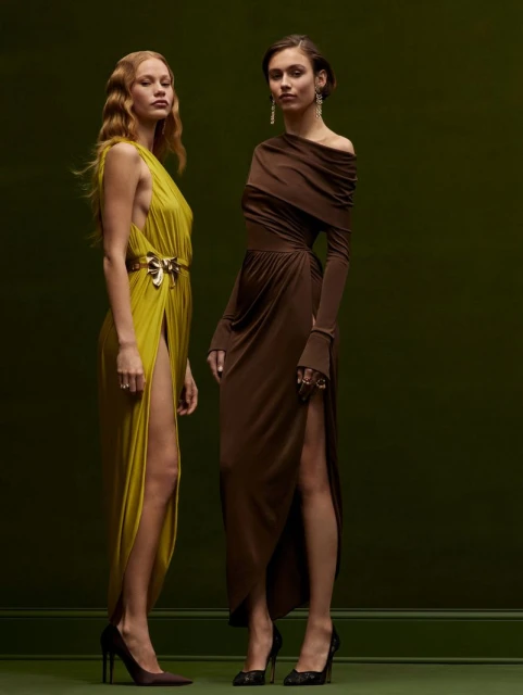 Two women in draped evening gowns, one in chartreuse with a gold belt, the other in brown, pose against a dark green background.