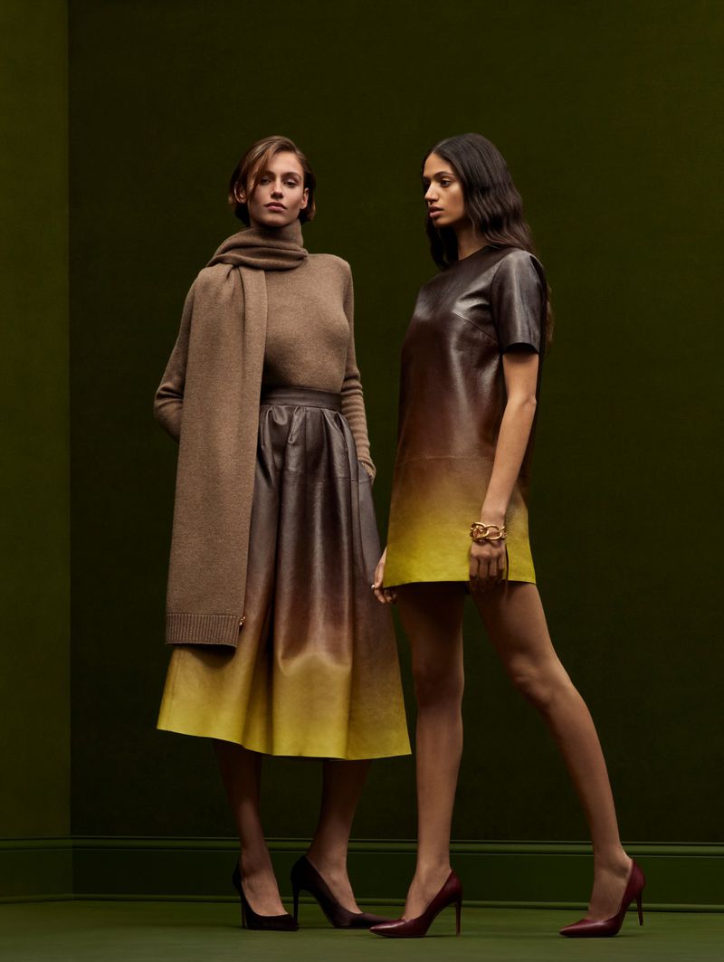 Two women in a fashion studio, one wearing a brown sweater and skirt with a scarf, the other in a brown-and-yellow dress.