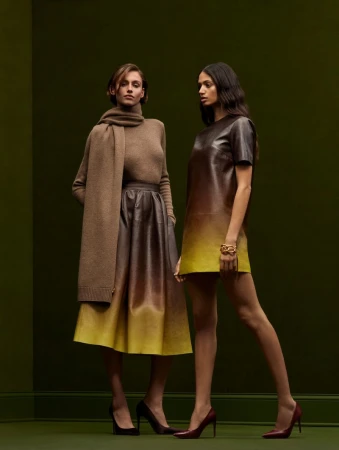Two women in a fashion studio, one wearing a brown sweater and skirt with a scarf, the other in a brown-and-yellow dress.