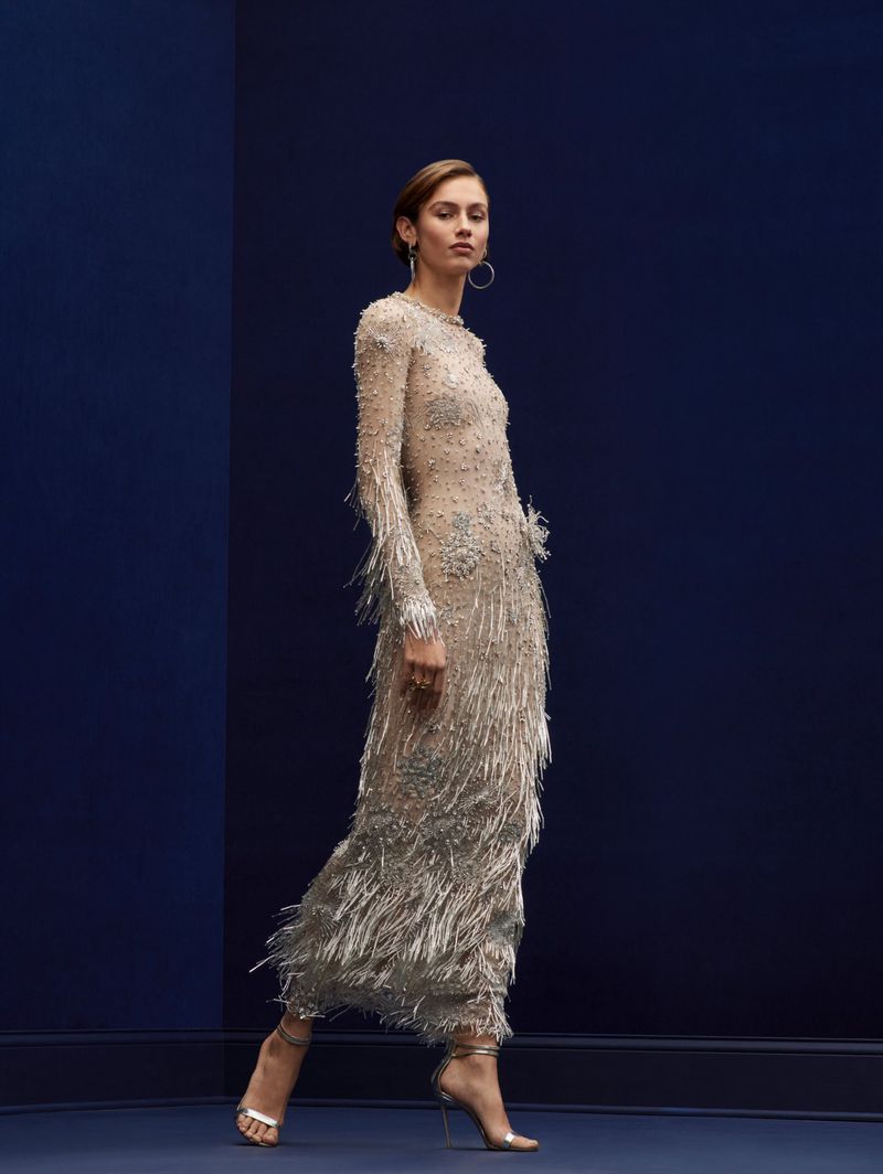 Woman walks in full-length beaded dress with fringe detail and heeled sandals against a navy blue wall in a fashion studio.