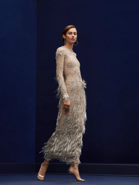 Woman walks in full-length beaded dress with fringe detail and heeled sandals against a navy blue wall in a fashion studio.
