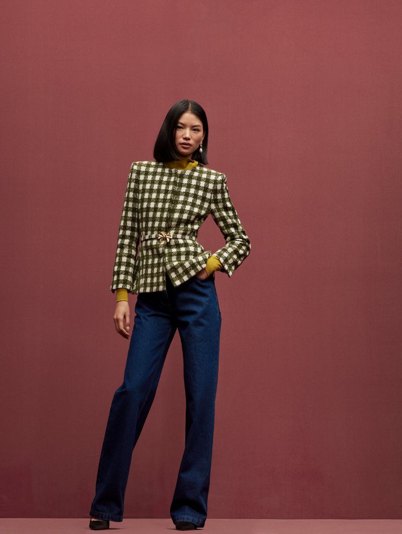 A full-length studio portrait of an Asian woman in a yellow turtleneck, plaid blazer with a belt, and blue jeans against a pink background.